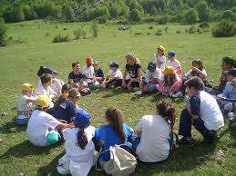 The image depicts a group of children sitting in a circle on the grass, with a woman standing in the center, likely engaged in an outdoor activity or educational session.