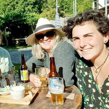 Das Bild zeigt zwei Frauen, die an einem Tisch mit Getränken und Essen sitzen, umgeben von einer ruhigen Umgebung mit Bäumen und einem Innenhof im Hintergrund.