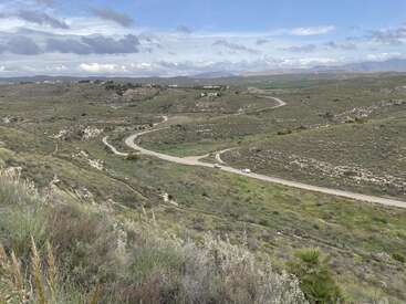 Eine kurvenreiche Straße schlängelt sich durch eine grüne, hügelige Landschaft unter einem teilweise bewölkten Himmel. Die spärliche Vegetation und die Berge in der Ferne schaffen eine ruhige, offene ländliche Atmosphäre.