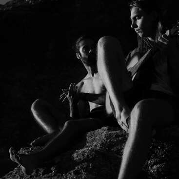Zwei Männer sitzen in dramatischer Beleuchtung auf Felsen. Einer spielt konzentriert Gitarre, während der andere ausdrucksstark gestikuliert. Die Szene strahlt Intimität, Kreativität und eine entspannte Atmosphäre aus.