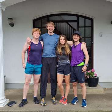 Vier Freunde in Sportkleidung stehen Arm in Arm und lächeln auf einer Veranda vor einer Tür. Neben ihnen stehen Topfblumen, die auf ein freundliches, zwangloses Beisammensein hindeuten.