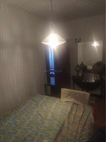 Ein kleines, schwach beleuchtetes Schlafzimmer mit einer gemusterten Tagesdecke, einer hängenden Deckenlampe, einem Spiegel, einem Waschtisch und einer teilweise offenen Tür. Gemütliche Atmosphäre.