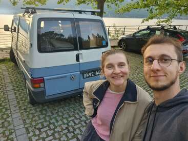 La imagen muestra a una joven pareja posando para un selfie delante de una furgoneta Volkswagen y otros coches, con una vista panorámica de una masa de agua al fondo.