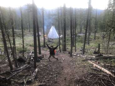 Eine Person steht aufgeregt mit erhobenen Armen auf einer Waldlichtung. Im Hintergrund sind zwischen hohen Bäumen und Bergen ein Tipi-ähnliches Zelt und ein kleines Lagerfeuer zu sehen.