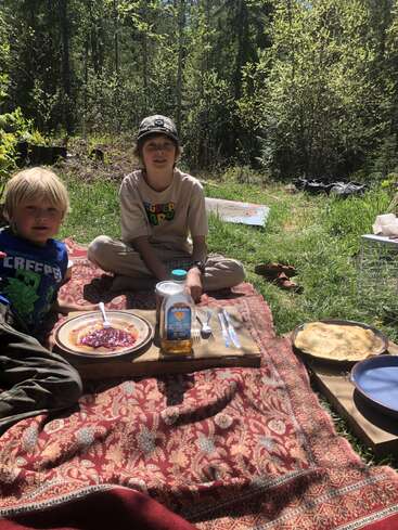 Zwei Kinder genießen ein Picknick im Freien auf einer gemusterten Decke, umgeben von grünem Wald. Pfannkuchen, Marmelade, Honig und Teller stehen bereit für ein sonniges, gemütliches Essen.