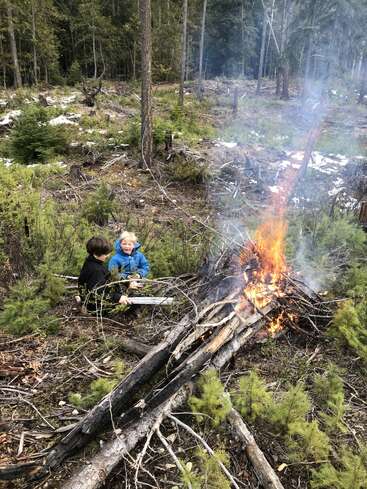 Zwei Kinder sitzen an einem kleinen Lagerfeuer auf einer Waldlichtung. Umgestürzte Äste brennen, Rauch steigt auf, und auf dem Boden liegen Zweige, Grünzeug und Schneeflecken verstreut.