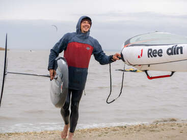 A smiling man in a wetsuit and hoodie stands on a beach, holding a foil surfboard and an inflatable board beside the water on a cloudy day.