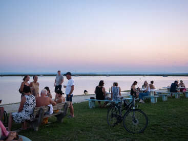 People gather by a serene lakeside at sunset, sitting on benches and picnic tables, chatting, relaxing, and enjoying the view. A bicycle is parked nearby.