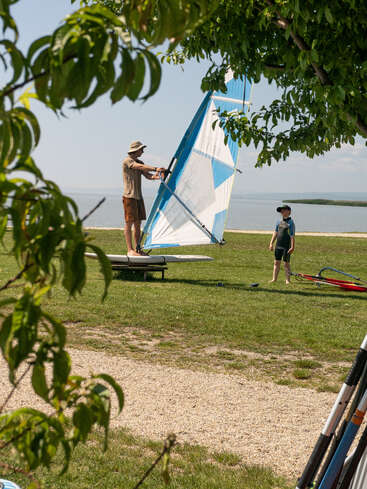 A man demonstrates windsurfing on land while a child watches nearby. The scene is framed by tree branches, with a shoreline and calm water in the background.