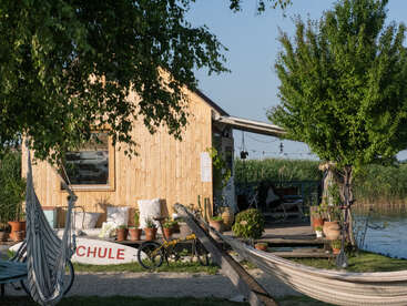 A cozy wooden cabin sits by a river, surrounded by potted plants, hammocks, bicycles, and trees. Peaceful, sunny setting evokes relaxation and nature’s tranquility.