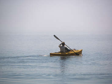 A person paddles a yellow kayak across calm, serene water under a hazy sky, creating gentle ripples as they move away, embodying peaceful solitude and adventure.