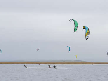 Several people are kite surfing in the water under a cloudy sky. Brightly colored kites soar overhead, while a sandy embankment stretches across the background.