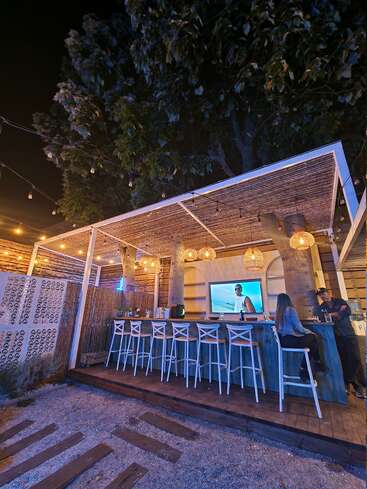 L'image représente un bar avec une terrasse en bois et un toit blanc, avec un écran de télévision, des tabourets de bar et un homme debout derrière le bar.