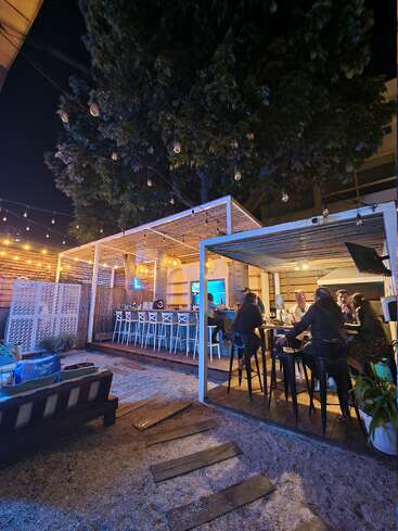 L'image représente un bar avec un espace extérieur couvert, comprenant un bar, des tabourets et des tables, entouré d'un sol en gravier et d'un arbre en arrière-plan.