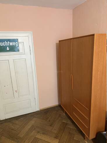 Uma porta branca com uma placa alemã “Fluchtweg” (rota de fuga) está em uma parede rosa ao lado de um guarda-roupa de madeira sobre um piso de madeira em zigue-zague.