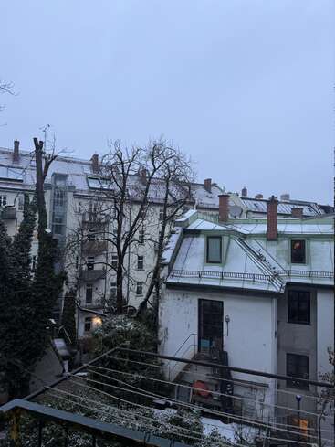 Uma cena urbana de inverno com telhados e árvores cobertos de neve. Prédios altos cercam um pequeno pátio. O céu está nublado, criando uma atmosfera fria e tranquila.
