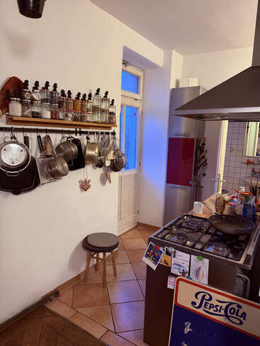 Essa cozinha aconchegante tem panelas, frigideiras e utensílios pendurados, potes de temperos numa prateleira, um banquinho, fogão a gás, geladeira e um letreiro retrô da Pepsi-Cola. Iluminação aconchegante.