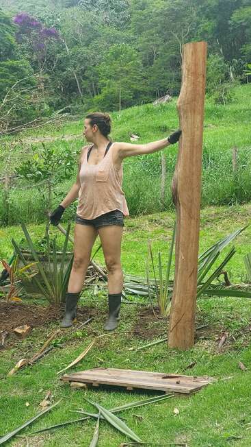 A woman stands outdoors in a lush, green garden holding a large wooden post. She wears gloves, boots, shorts, and a tank top, surrounded by plants and debris.