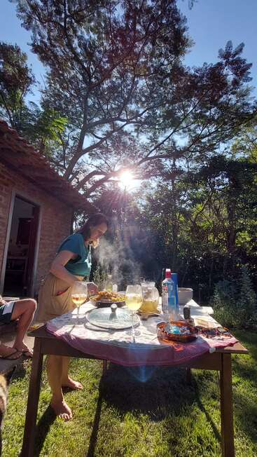 A woman prepares food at a sunlit outdoor table, surrounded by trees and drinks, with steam rising and nature creating a peaceful, warm and inviting atmosphere.