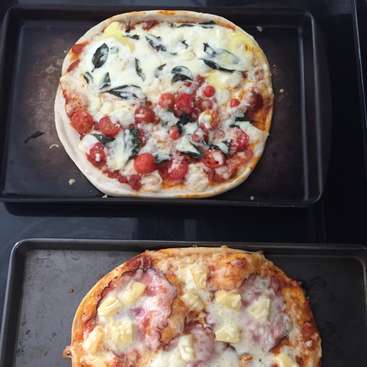 The image shows two pizzas on baking trays, one with tomatoes and basil, the other with ham and pineapple, both topped with melted cheese.