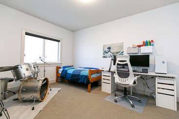 This tidy bedroom features a drum set, single bed with blue bedding, a modern desk setup with computer, ergonomic chair, and minimalist decor with wall art.