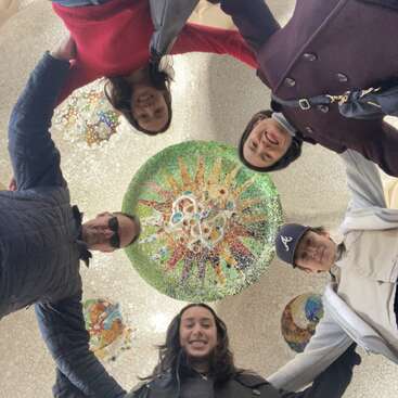 The image shows a group of five people standing in a circle, looking down at the camera, with a mosaic sun design on the ground below them.