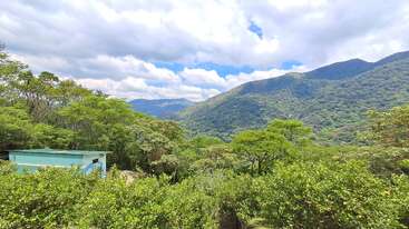 Lush green trees cover the hillside landscape under a vibrant blue sky. A small turquoise building sits nestled among the foliage. Rolling mountains dominate the background.