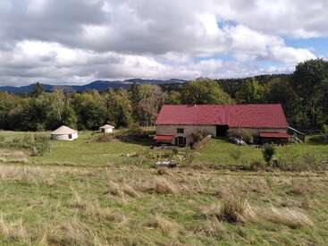 A rustic farmhouse with a red roof sits among greenery and trees, two white yurts nearby, under a cloudy sky, surrounded by mountains and grassy fields.