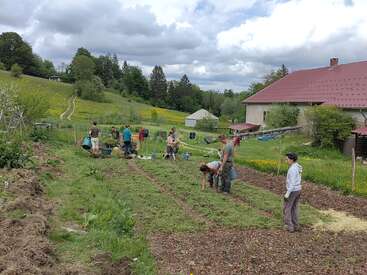 A group of people are working together in a large garden, surrounded by green fields, trees, and a farmhouse with a red roof.