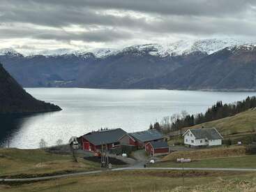 Un sereno paisaje de fiordos presenta graneros rojos y una casa blanca sobre césped en pendiente, con montañas nevadas y aguas tranquilas bajo un cielo nublado en Noruega.