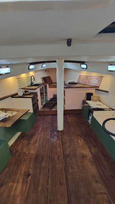 This image shows the interior of a boat cabin, featuring wooden floors, a kitchenette area, small windows, and various surfaces with partially completed construction or remodeling.