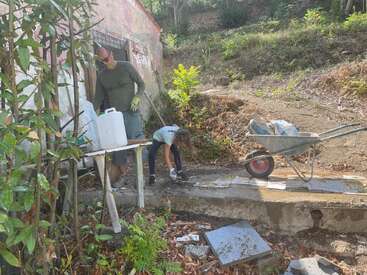 Un hombre y un niño están trabajando al aire libre cerca de un edificio rústico, utilizando guantes, una carretilla y jarras, rodeados de plantas y materiales de construcción en un día soleado.