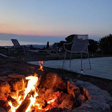 A glowing campfire burns brightly in the foreground, surrounded by stones, with empty chairs and loungers overlooking a tranquil sunset view by the sea.