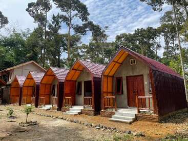 Cuatro pequeñas cabañas de madera con tejados rojos están situadas una al lado de la otra en un bosque, rodeadas de árboles altos, bajo un cielo parcialmente nublado. Ambiente tranquilo.