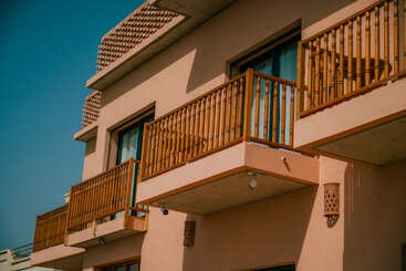 Cette image montre un bâtiment moderne avec des balcons en bois, des murs beiges, de grandes fenêtres et des lumières extérieures, sous un ciel bleu lumineux.