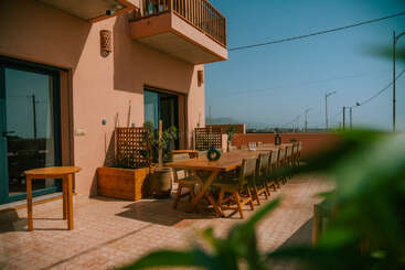 Un patio extérieur ensoleillé est doté d'une longue table à manger en bois avec des chaises, des plantes en pot, des murs couleur pêche et des portes coulissantes bleues, créant ainsi une atmosphère accueillante.