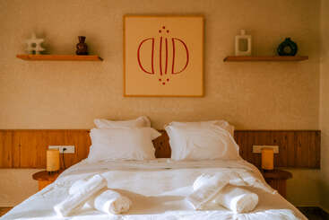 Un lit double bien fait avec des draps et des oreillers blancs, deux serviettes roulées, des tables d'appoint en bois, une décoration moderne, des étagères et un tableau d'art minimaliste au-dessus de la tête de lit.