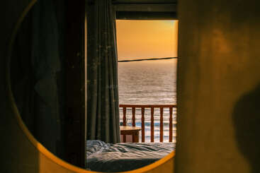 Une chambre à coucher confortable se reflétant dans un miroir rond, donnant sur un balcon avec des balustrades en bois et un magnifique coucher de soleil sur l'océan, évoquant la chaleur, la paix et la relaxation.