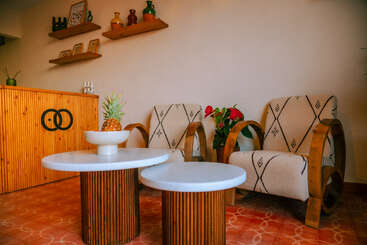 Cette pièce confortable comprend deux fauteuils à motifs, des tables rondes en bois avec des plateaux blancs, un bol avec un ananas, des étagères décoratives et un bureau d'accueil en bois.