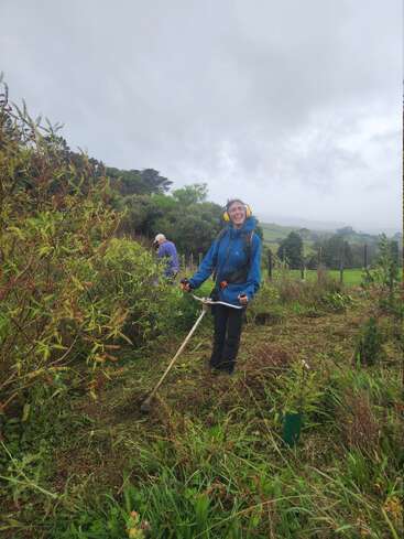 Una persona sonriente con una chaqueta azul utiliza una desbrozadora al aire libre, rodeada de vegetación y bajo un cielo nublado. En el fondo, otra persona trabaja cuidando las plantas.