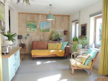 This cozy living room features a brown sofa, pastel cushions, wooden wall art, abundant houseplants, large windows, a comfy armchair, and plenty of natural sunlight.