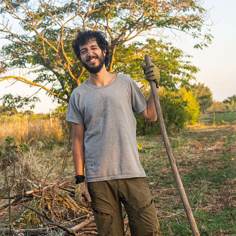 Um homem sorridente com cabelos cacheados, vestindo uma camiseta cinza e luvas, está ao ar livre segurando uma pá, cercado por árvores e pela luz do sol em um ambiente natural.