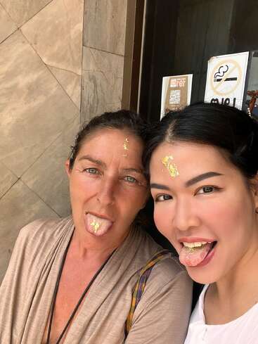 Two women pose for a selfie, smiling with their tongues out. Both have shiny gold leaf on their foreheads and tongues, showing playful, joyful expressions.