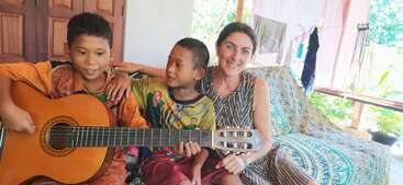 A imagem mostra uma mulher e um menino sentados juntos em um sofá, com o menino segurando um violão e a mulher sorrindo para a câmera.