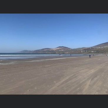 A imagem retrata uma cena de praia serena com uma vasta extensão de areia, ondas suaves e uma cadeia de montanhas distante sob um céu azul claro em um dia ensolarado.