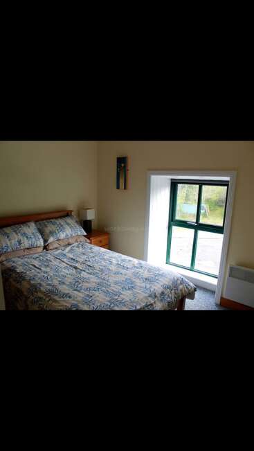 A imagem mostra um quarto sereno com uma cama de casal, cabeceira de madeira e mesa de cabeceira, com uma janela com moldura verde e vista para o exterior.