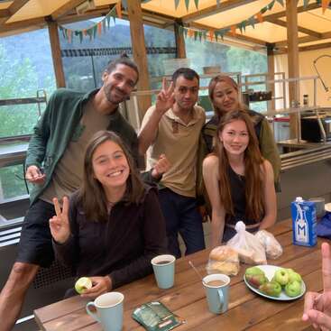 Das Bild zeigt eine Gruppe von sechs Personen, die für ein Selfie an einem Holztisch mit Essen und Getränken in einer Küche oder einem Essbereich mit schöner Aussicht posieren.