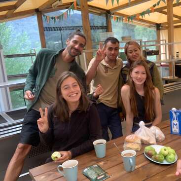 The image shows a group of six people posing for a selfie at a wooden table with food and drinks, in a kitchen or dining area with a scenic view.