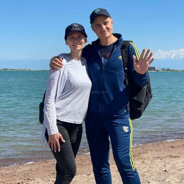 A smiling man and woman pose together on a sandy beach, with blue water and distant mountains in the background, wearing casual clothing and caps.
