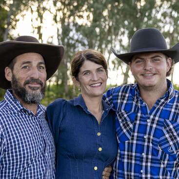Três adultos posam ao ar livre, sorrindo calorosamente. Dois homens usam chapéus de caubói e camisas xadrez, enquanto a mulher no meio usa um vestido azul. As árvores preenchem o plano de fundo.